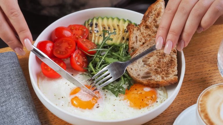 Close-up of a breakfast bowl with fried eggs, avocado, tomatoes, and bread, served with cappuccino.